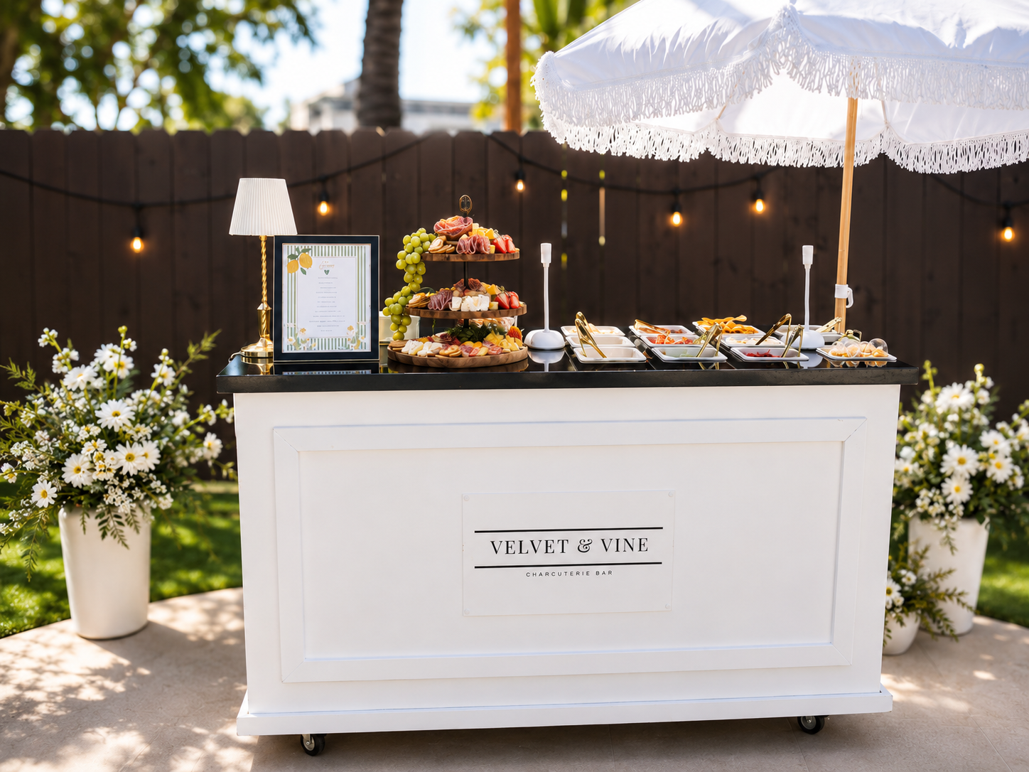 White Velvet & Vine charcuterie bar cart with tiered fruit and cheese display under a fringed umbrella